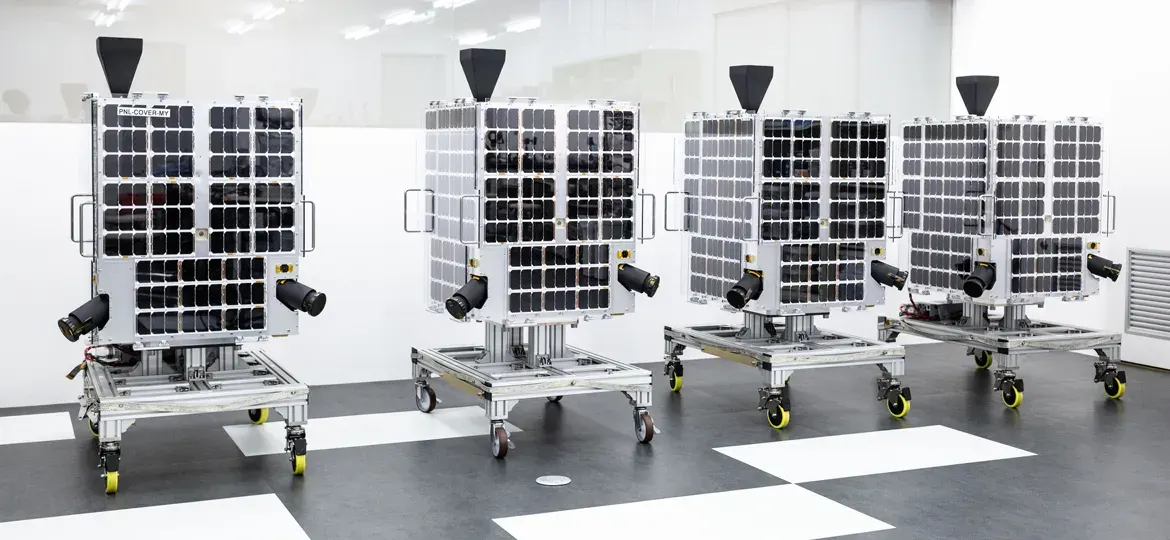 Row of cube satellites with solar panels mounted on wheeled platforms inside a clean indoor facility