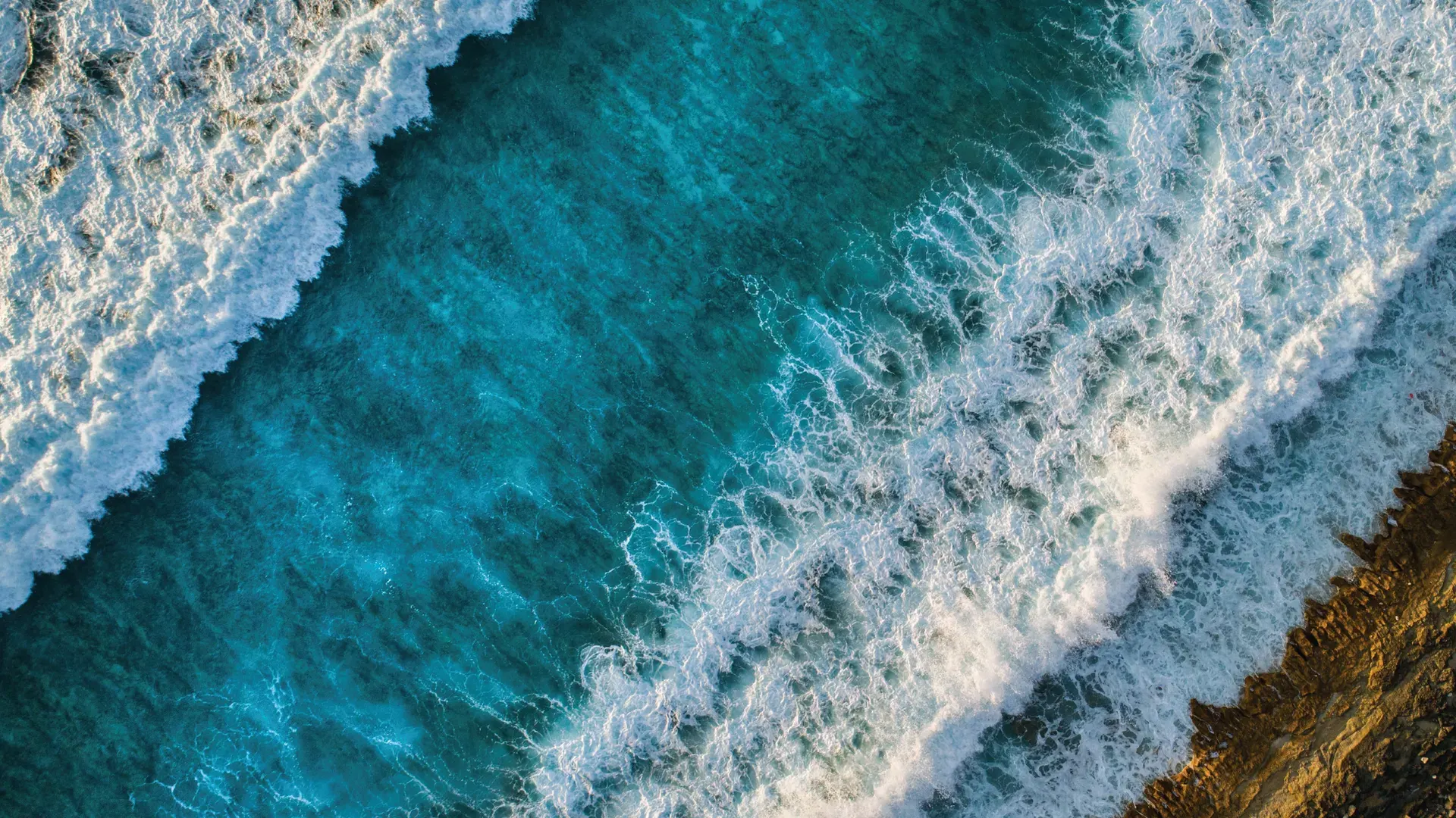 Aerial view of ocean waves crashing against a rocky shoreline with deep blue water and white foam