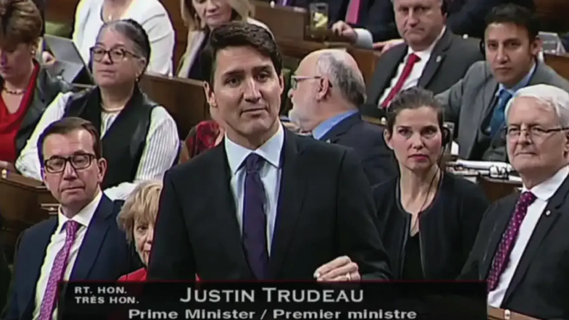Justin Trudeau speaking at a podium in a parliamentary chamber with seated officials around him